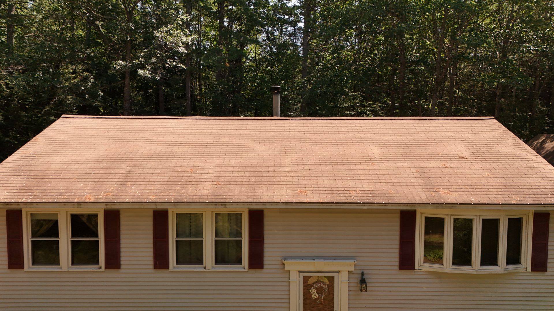 Beige house with maroon shutters and roof, surrounded by trees.