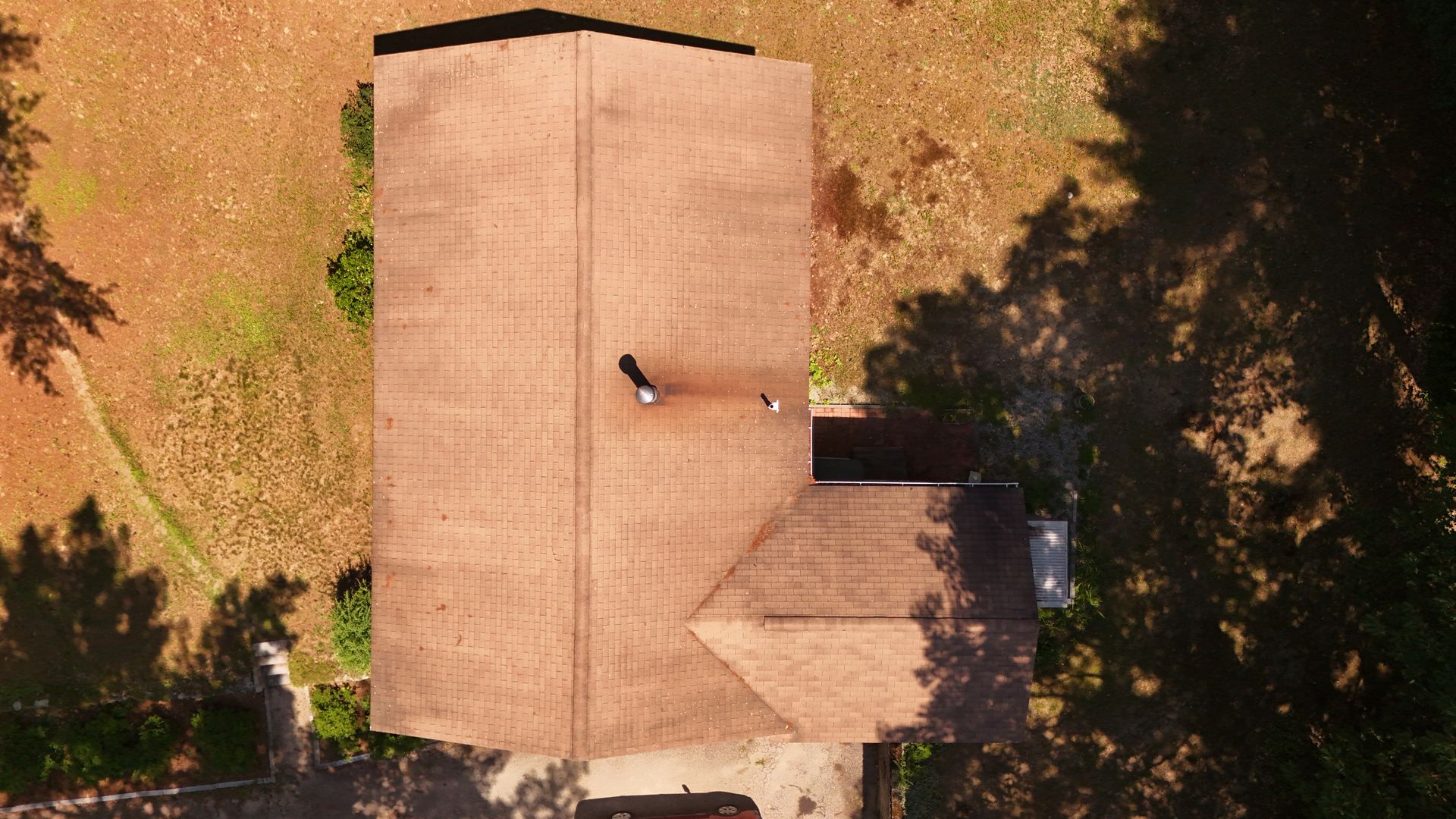 Overhead view of a brown-roofed house surrounded by brown and green foliage. Shadows from trees fall on the right side.