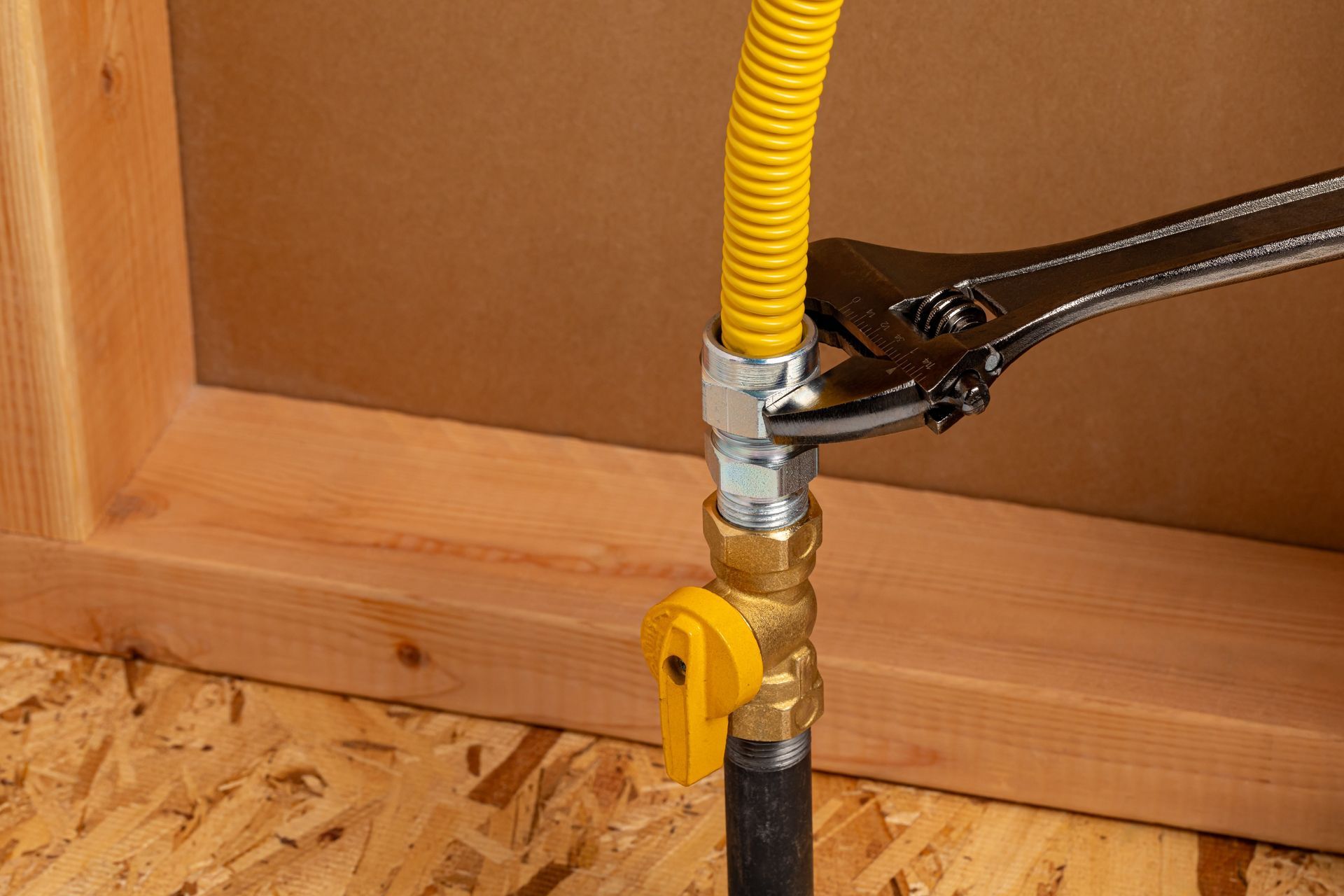 An adjustable wrench tightens a yellow corrugated gas line fitting onto a brass shut-off valve near a wooden wall.