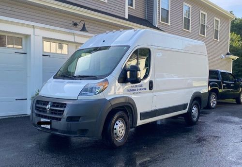 A white Ram ProMaster cargo van parked on an asphalt driveway in front of a house with a garage.