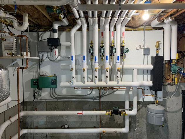Wall-mounted hydronic heating system with a manifold, multiple pumps, white piping, and a small expansion tank.