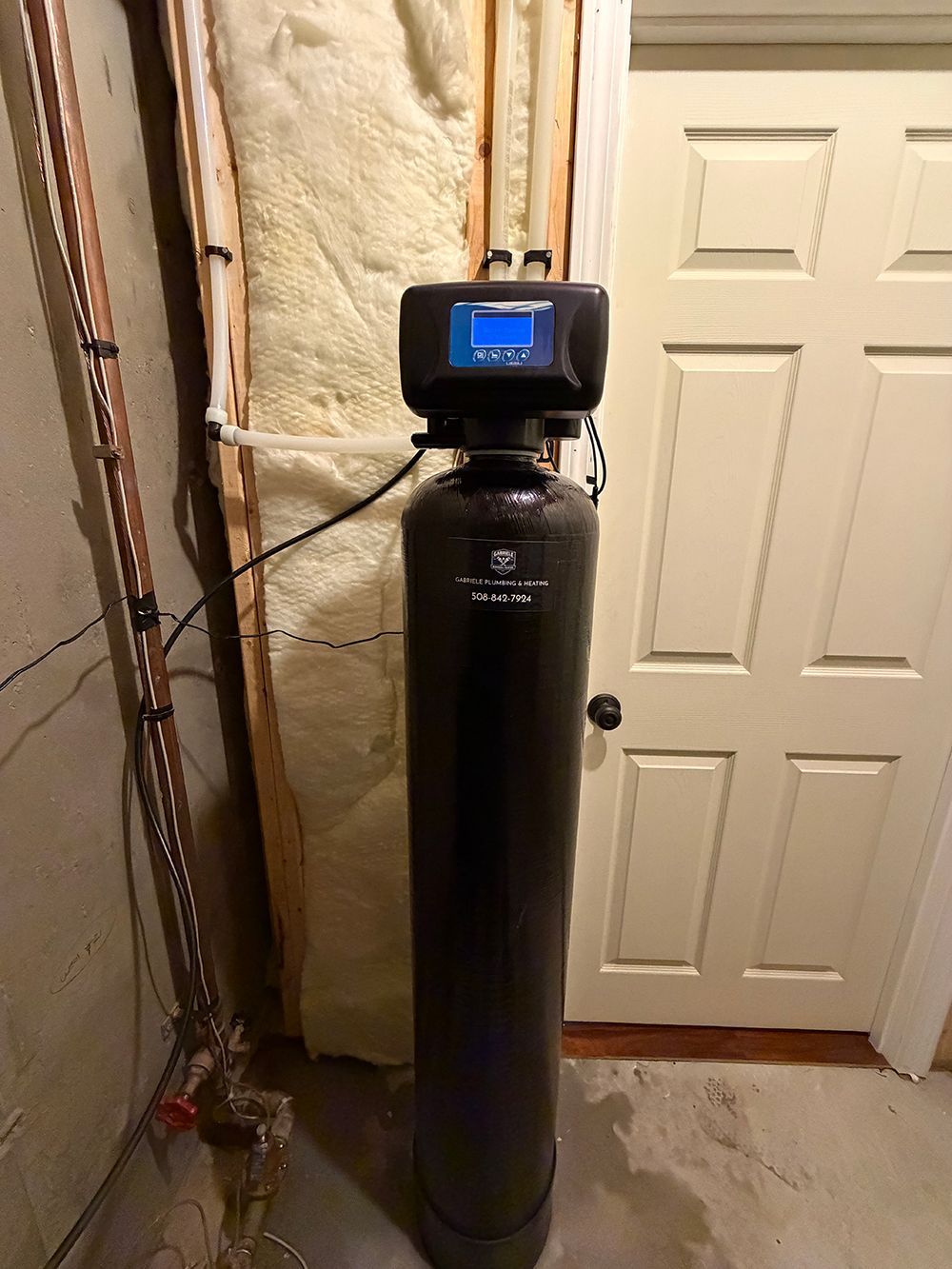 Water softener tank with digital control head in a narrow utility closet beside a paneled door.