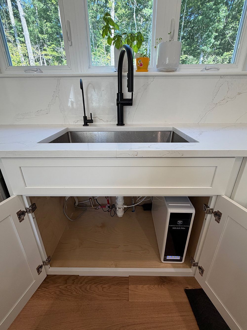 White kitchen sink with black faucet and open cabinet beneath, showing plumbing and a small appliance inside.