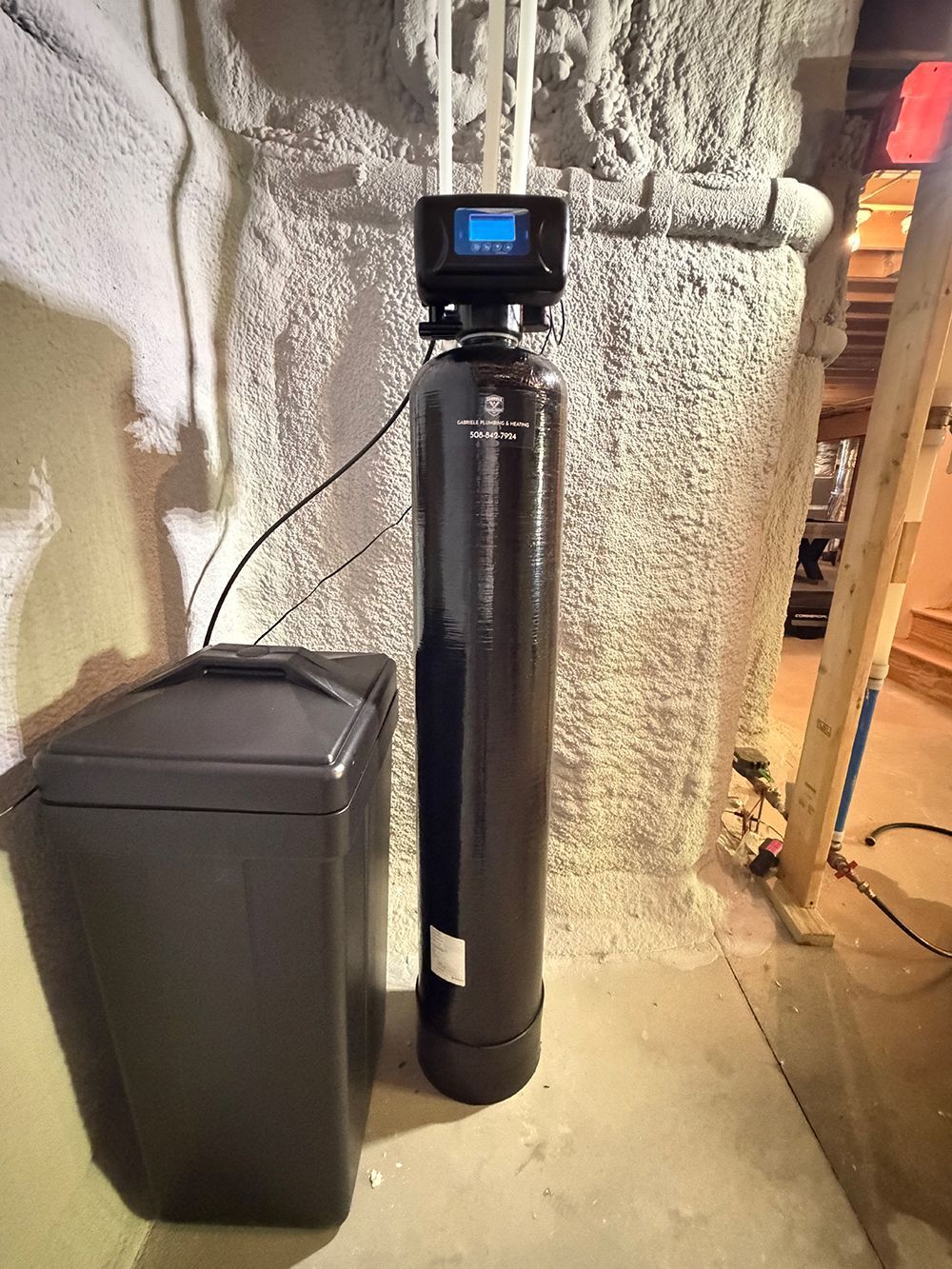 Black water softener tank with control head beside a dark trash bin in a basement utility area