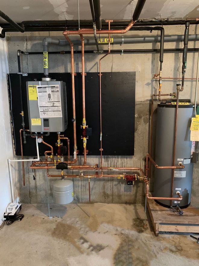 A wall-mounted tankless water heater connected to a storage tank with copper piping in a basement setting.