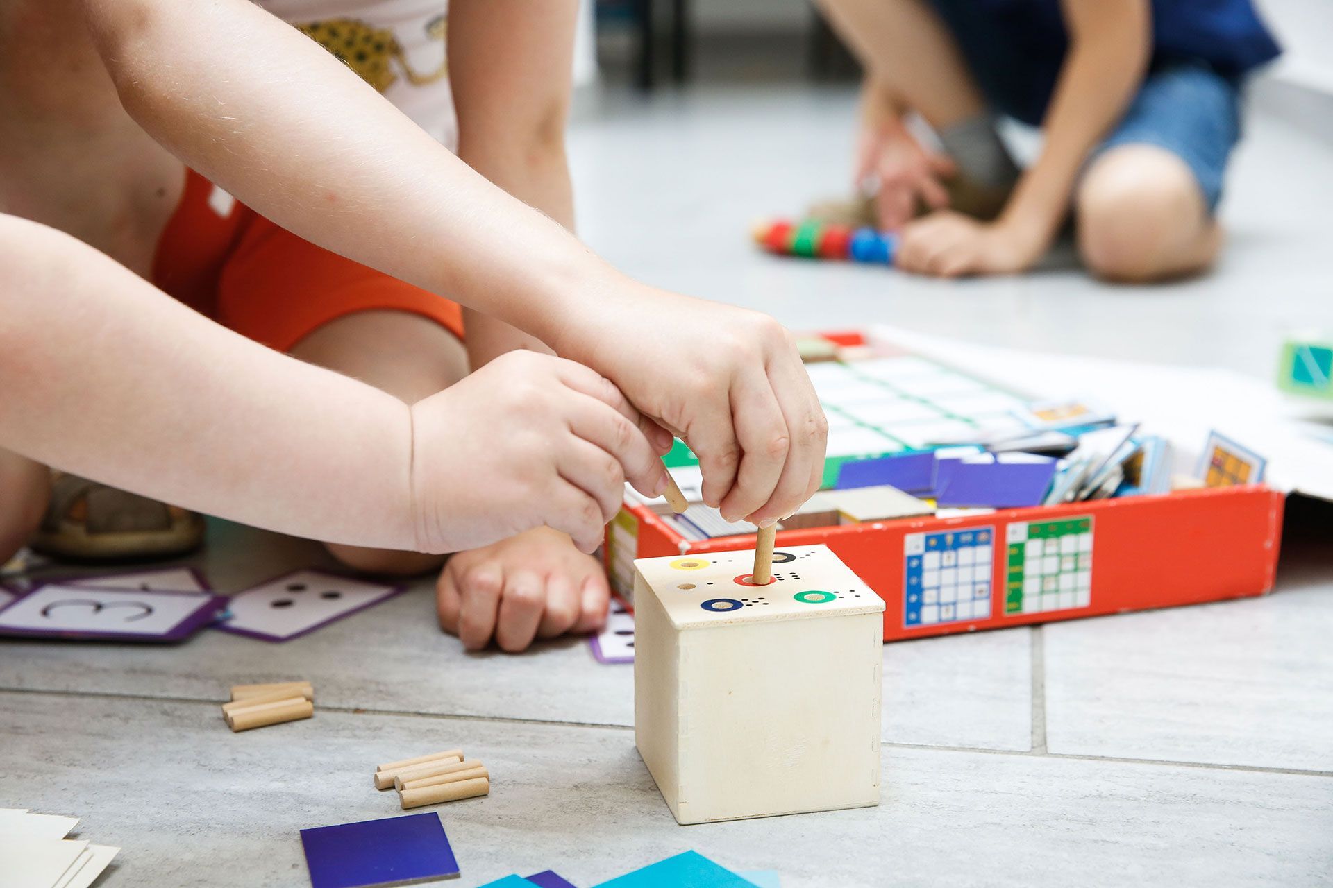 Children playing with a wooden block toy on a tiled floor.