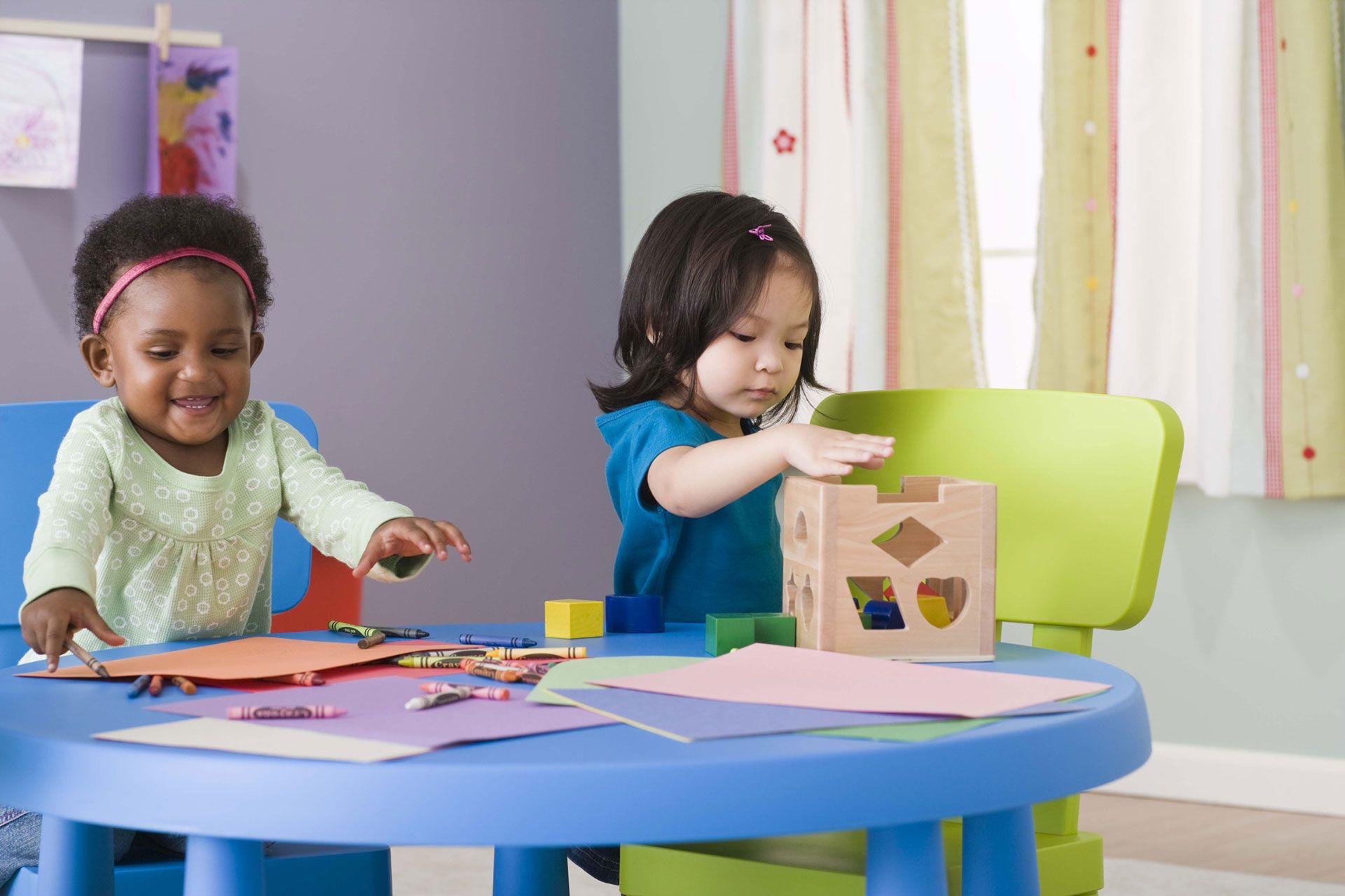 Two toddlers at a table, playing with crayons, paper, and a shape sorter.