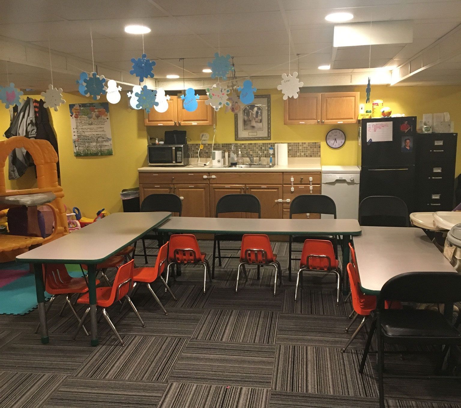 Classroom with tables, chairs, kitchenette, and play area, decorated with snowflakes.