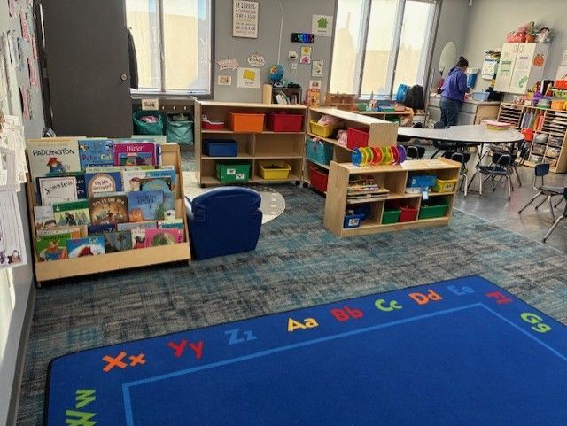 Classroom with bookshelves, colorful storage, blue rug with alphabet border, and a person near a table.