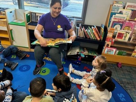 A woman reads a book to a group of children seated on a colorful rug.