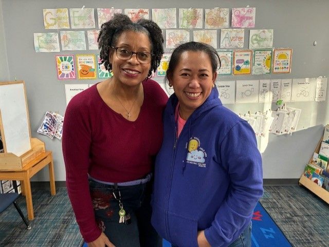 Two women smile, standing together in a classroom with children's artwork on the walls.