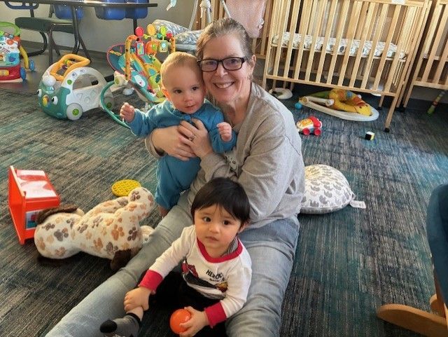 Woman smiles, holds one baby, another sits nearby. Babies, toys, cribs in a playroom.