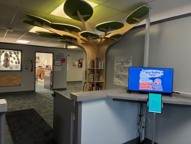 Library interior with a tree-shaped bookshelf, TV screen, and a counter.