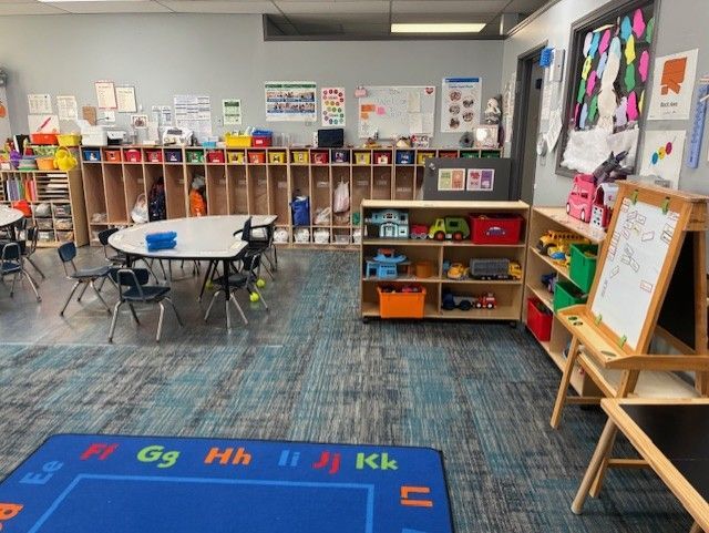 Preschool classroom with tables, chairs, cubbies, and toys. Blue and gray carpet, colorful displays, and a whiteboard.