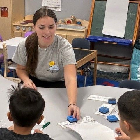 Teacher with two children at table, using blue stamps. Classroom setting. Smiling, engaged.