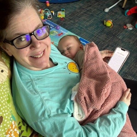 Woman holding sleeping baby, wearing glasses, blue shirt, pink blanket, phone in hand.