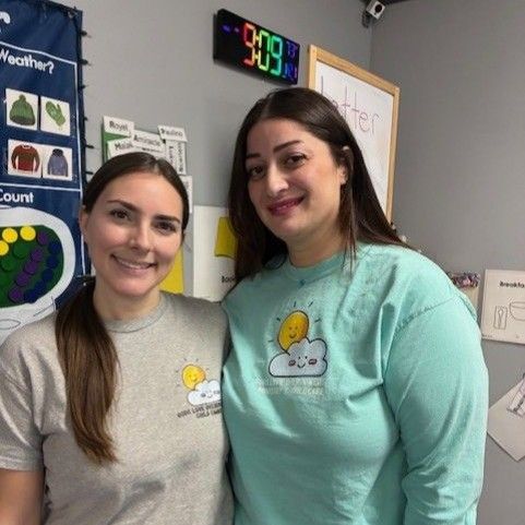 Two women smiling, wearing matching cloud logo shirts, in a classroom with educational displays.