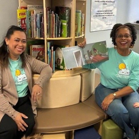 Two women in teal shirts sit near a bookshelf with books. One holds a book open, smiling.
