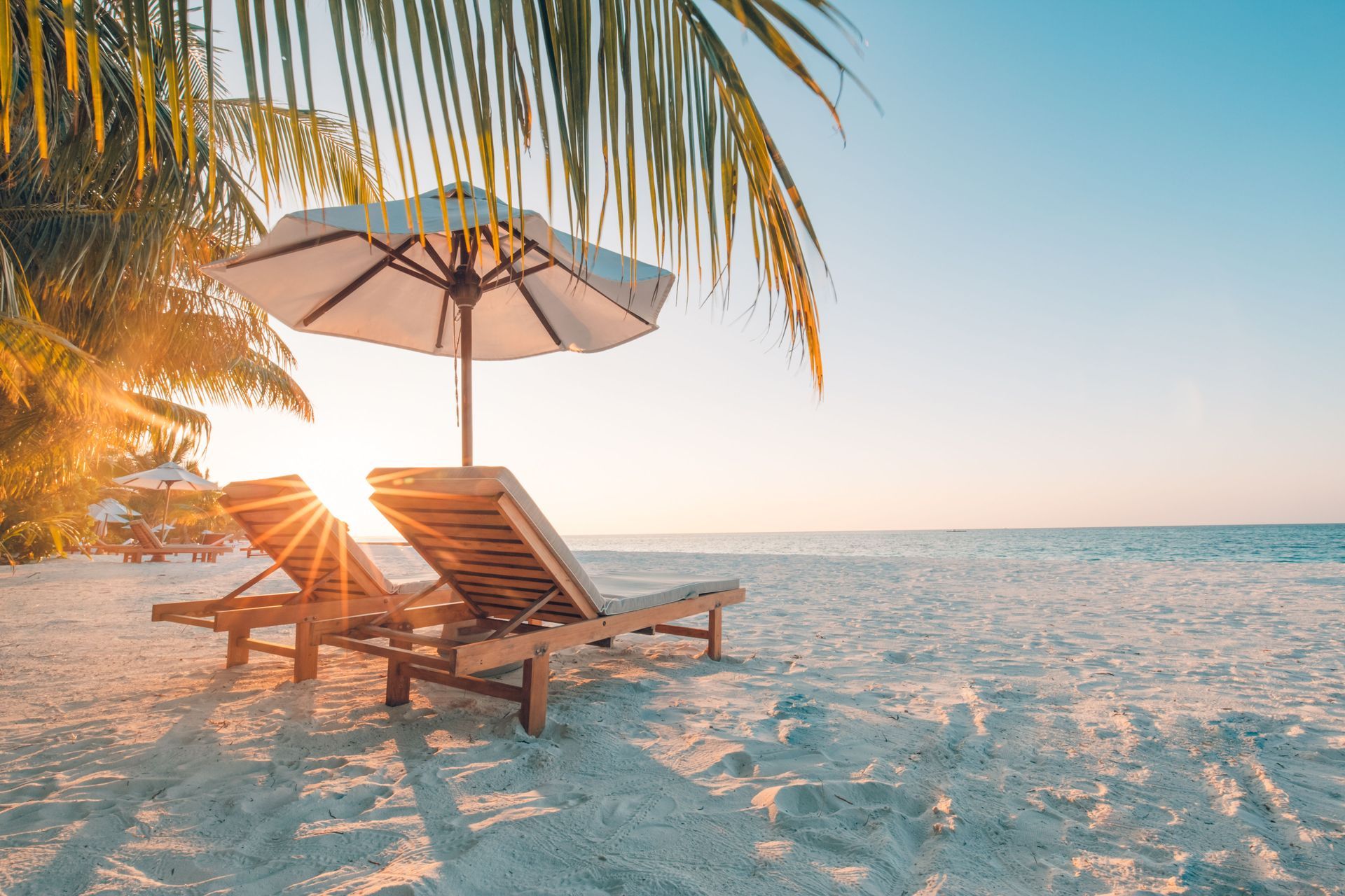 Two wooden lounge chairs under a white umbrella on a sandy beach at sunset.