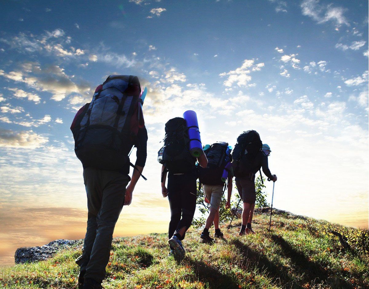 Hikers with backpacks climb a grassy hill under a partly cloudy sky.