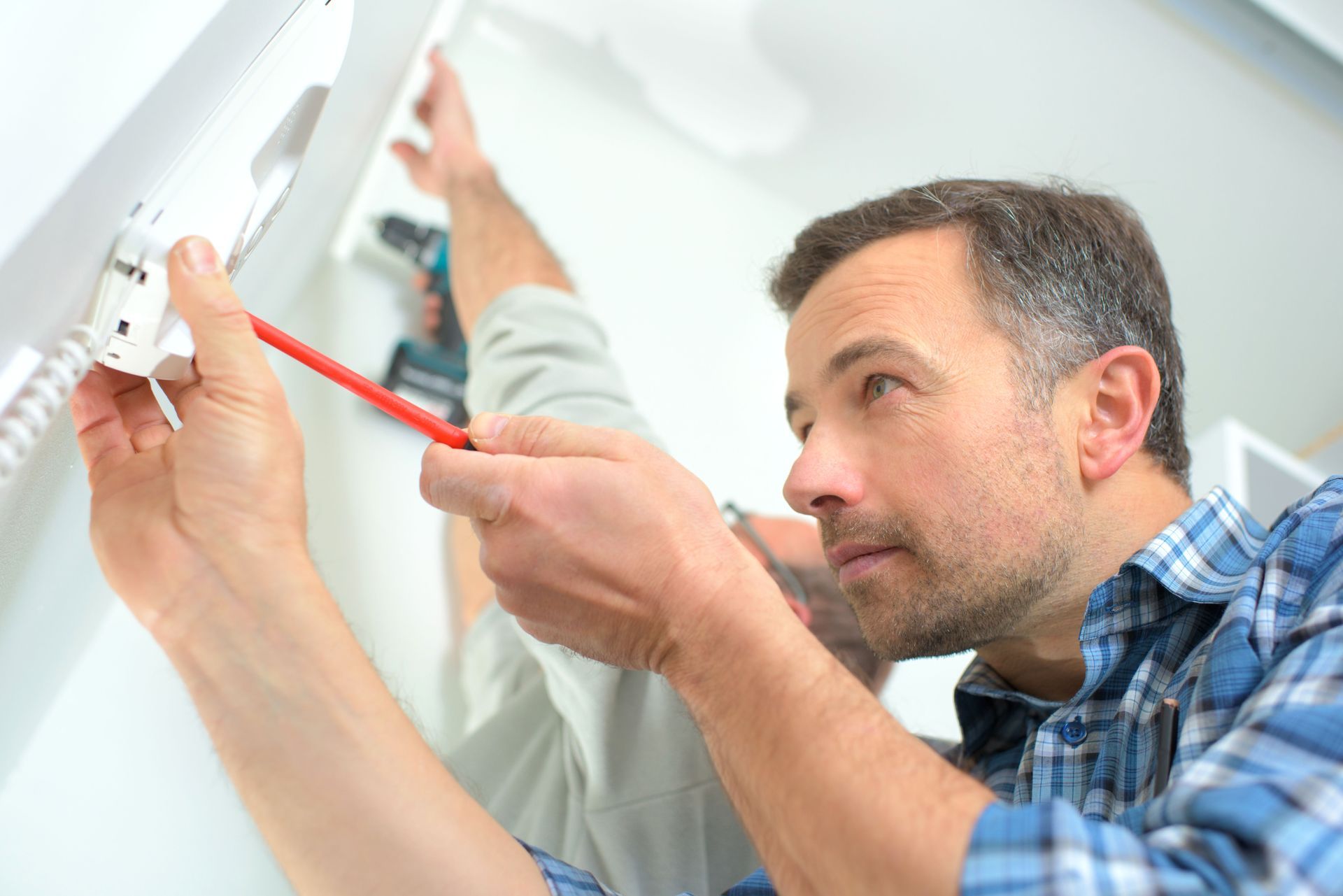Man using a screwdriver to install a wall-mounted smoke detector with another person assisting.