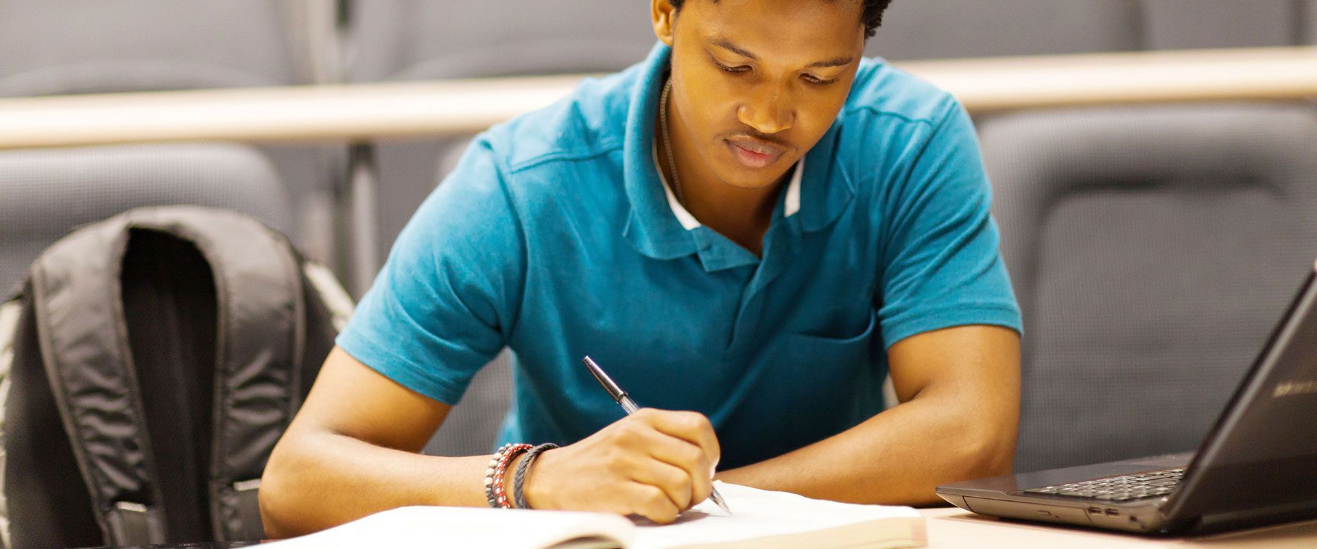 Person in a teal polo shirt writing in a notebook at a desk with a laptop and backpack in a classroom.