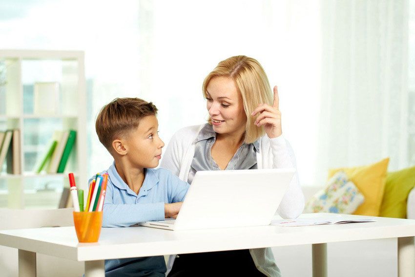Woman with laptop talking to child, both at a white desk in a room with a window and a bookshelf.