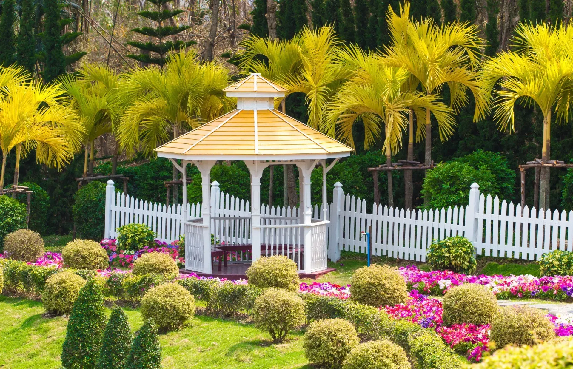 White gazebo in a colorful flower garden with palm trees and a white picket fence