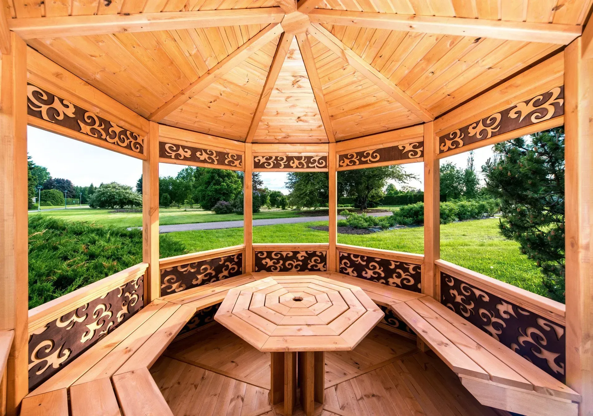 Wooden gazebo with carved benches overlooking a green field and trees