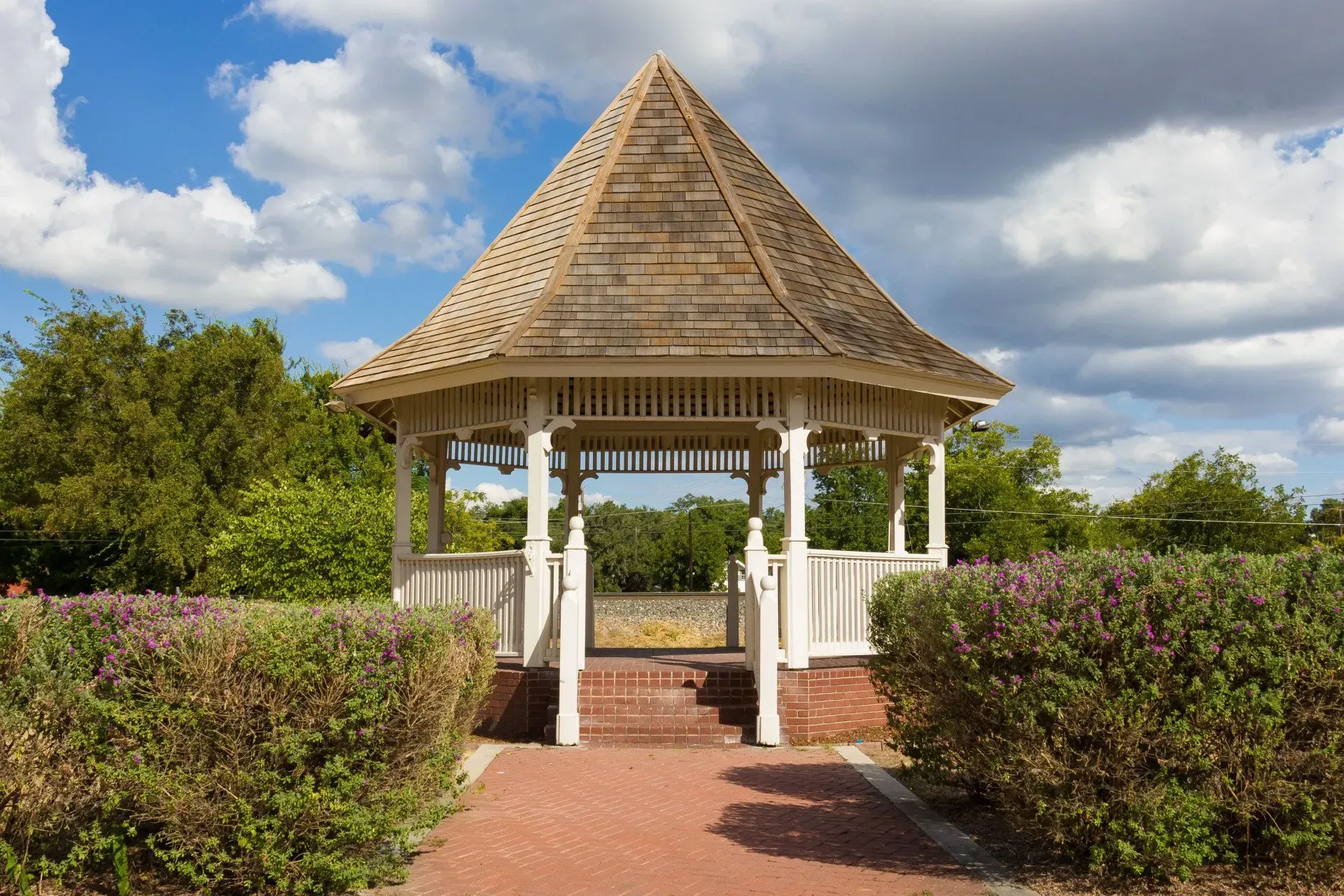 White gazebo with a thatched roof in a garden, framed by hedges under a cloudy sky