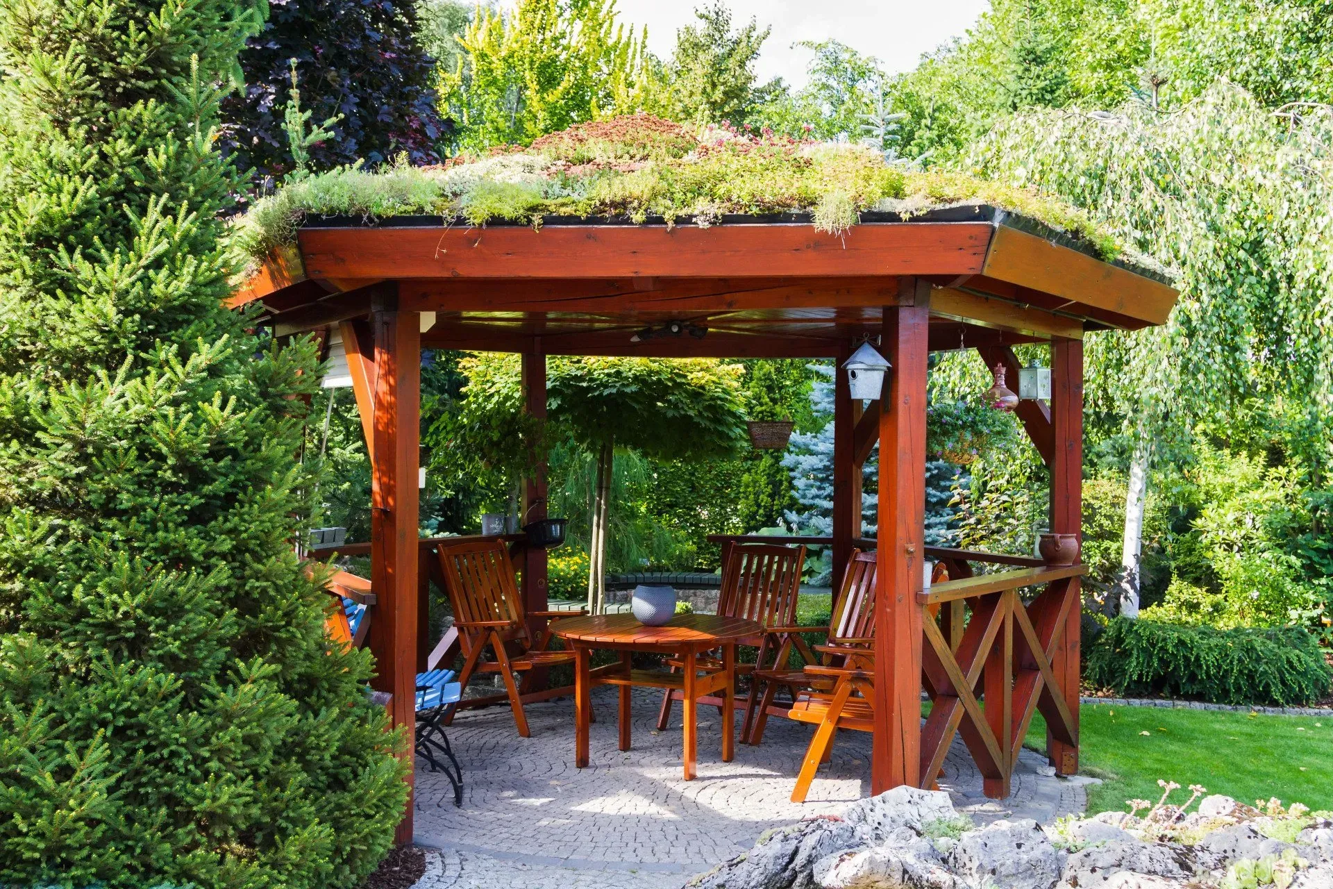 Wooden garden gazebo with chairs and table, surrounded by lush greenery and flowers
