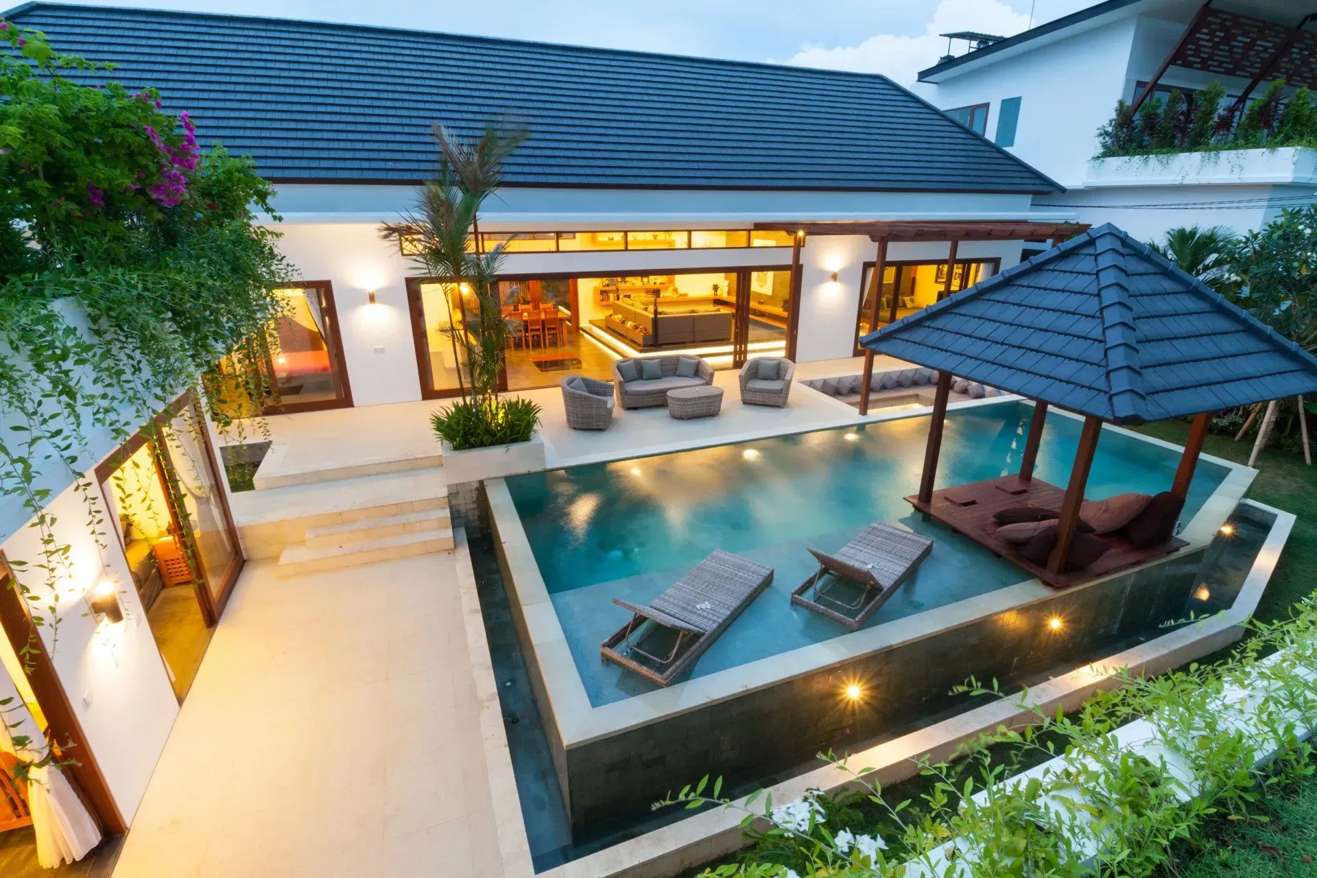 Modern luxury courtyard with a pool, lounge chairs, and a covered gazebo beside a lit villa at dusk