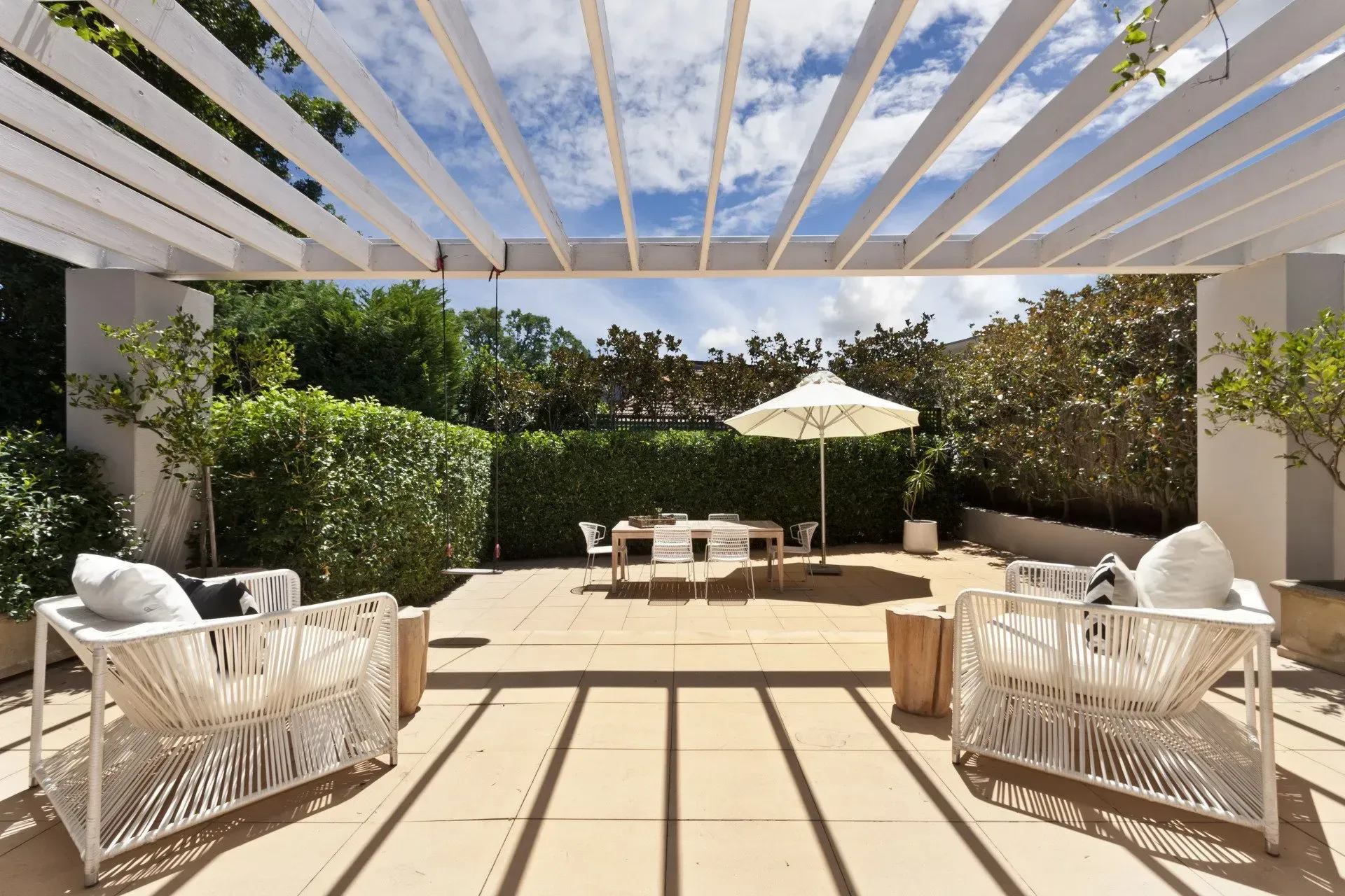 Sunlit patio with white pergola, wicker seating, and dining table overlooking lush greenery