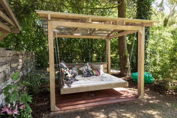 Wooden outdoor daybed under a pergola, with cushions in a leafy garden.