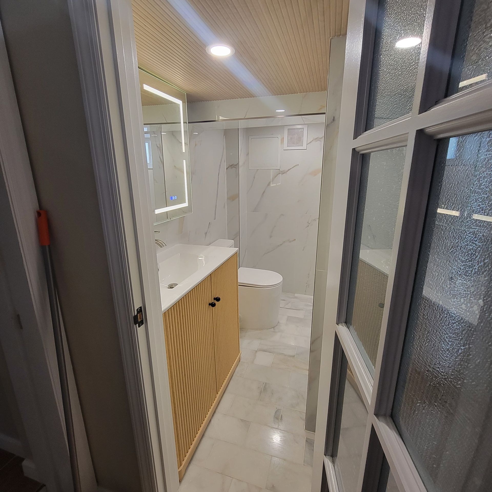 Bathroom with light wood vanity, marble tile, and a lit mirror.