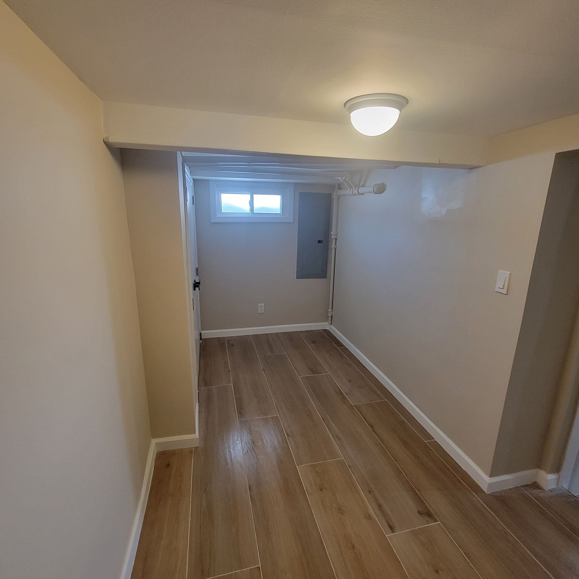Narrow room with wood-look floor, beige walls, and white trim. Ceiling light. Door and window at far end.