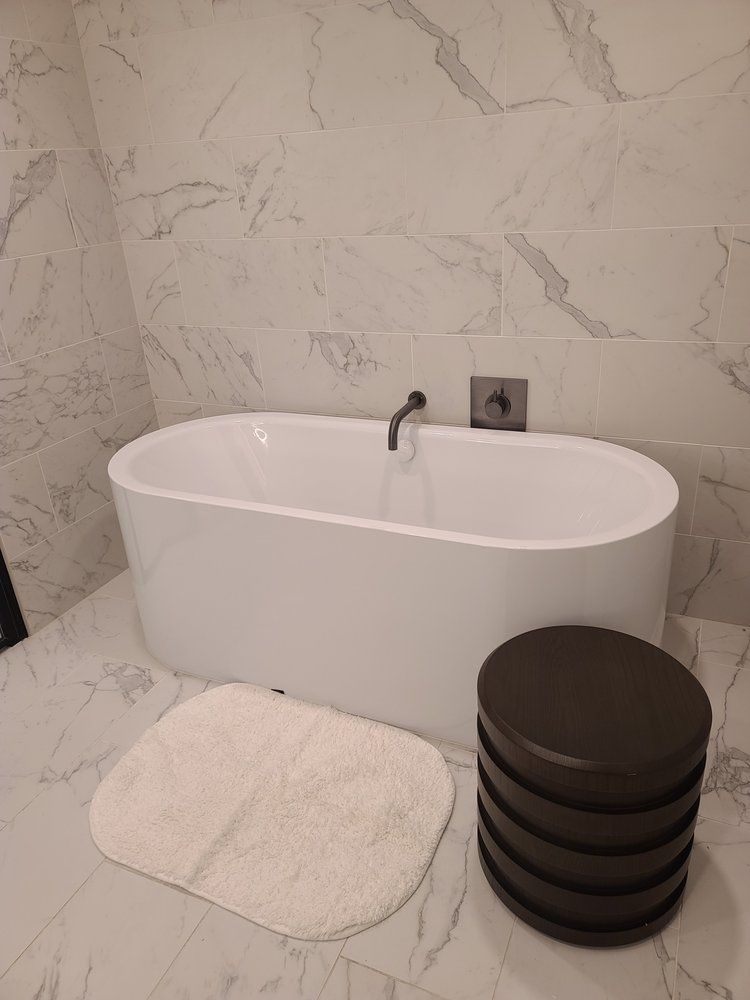 Oval bathtub in a modern bathroom with marble-look tiling. A dark wooden stool and a cream bath mat are nearby.