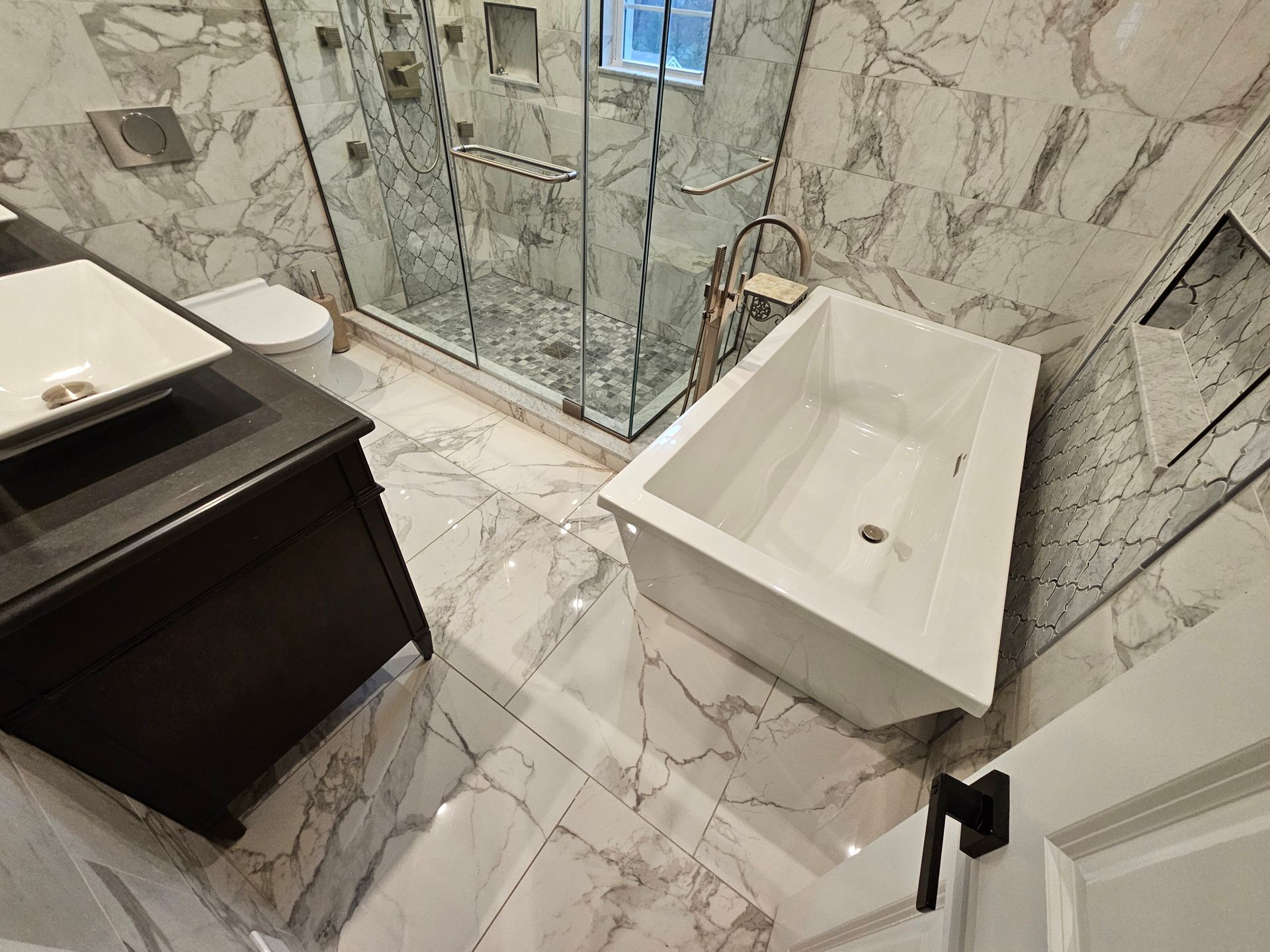 A luxurious bathroom with marble-patterned walls and floor. It features a white tub, glass shower, dark vanity, and toilet.