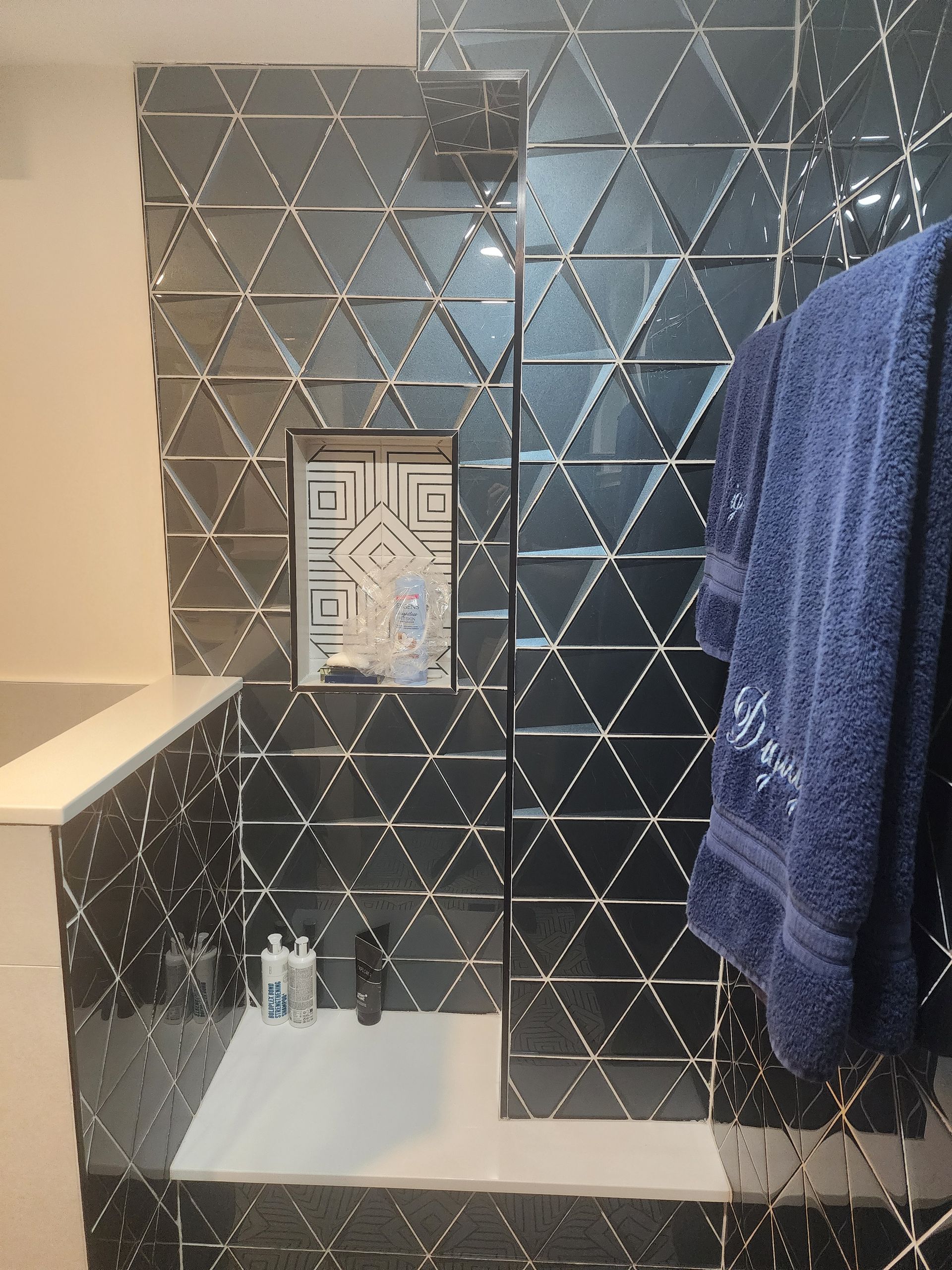 Black triangular tiles in a shower with a built-in shelf. A blue towel hangs on a bar, and toiletries sit on a ledge.