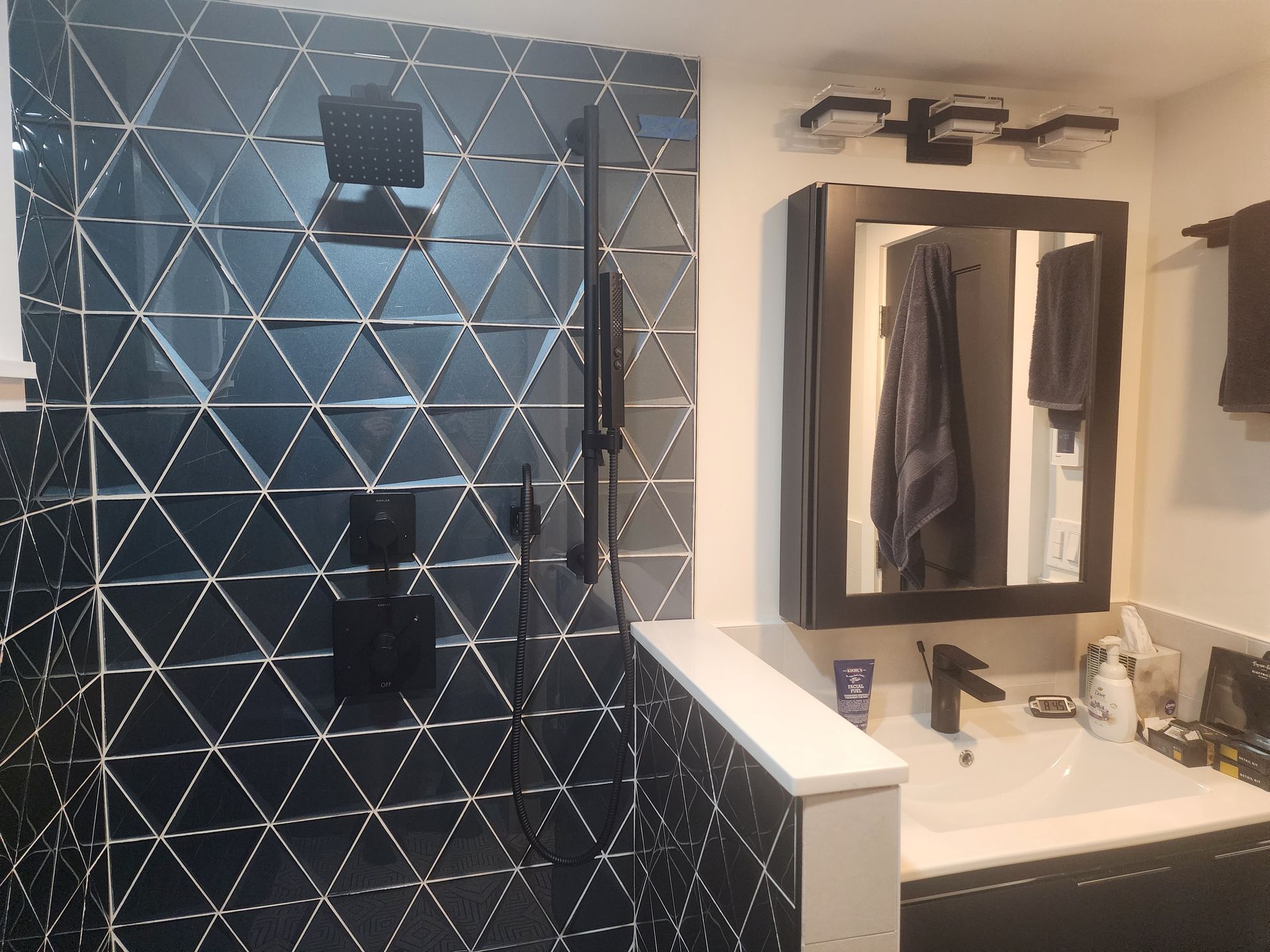 Bathroom with a black triangular-tiled shower, black fixtures, and a black vanity with a mirror and light fixture.