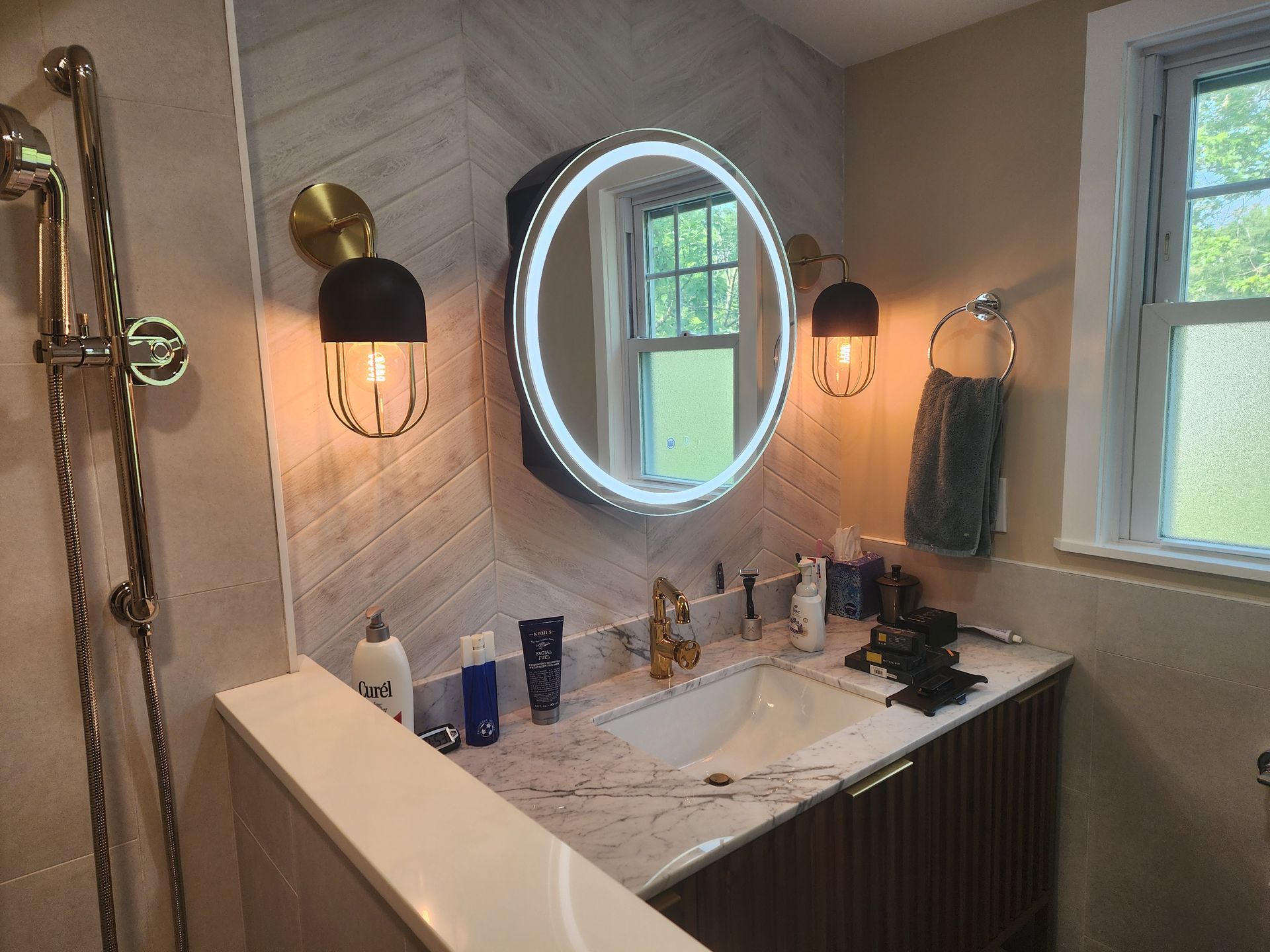 A modern bathroom with a white vanity, round mirror, and gold fixtures. The wall has a unique light-colored herringbone pattern.