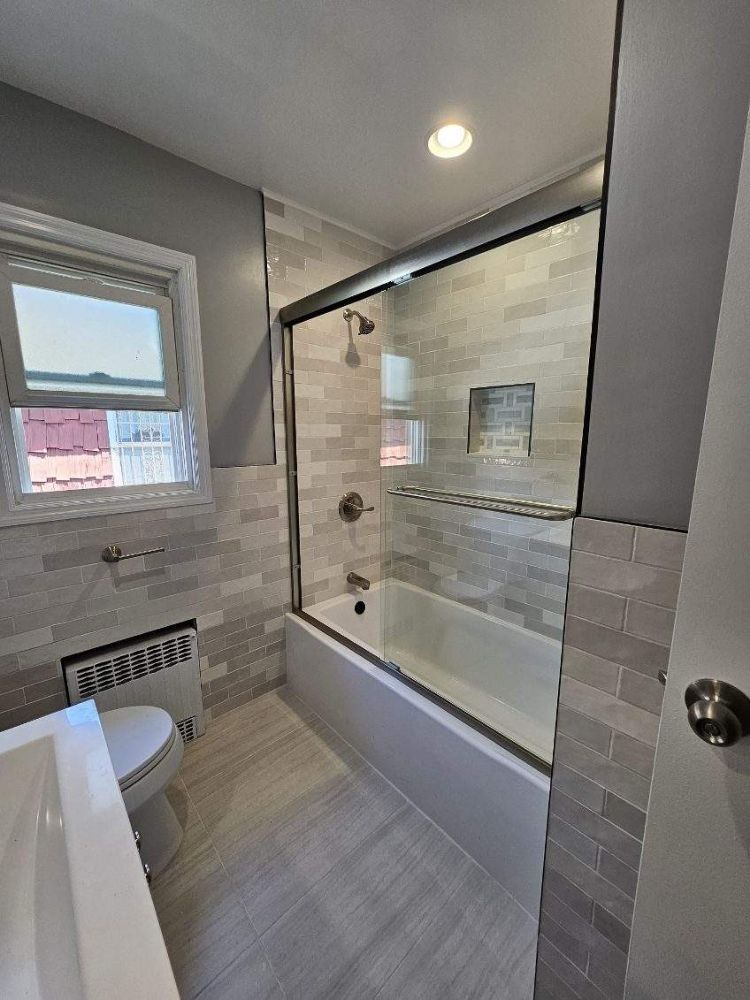 A renovated bathroom with light gray tile walls and flooring, a white tub and toilet, and a glass shower door.