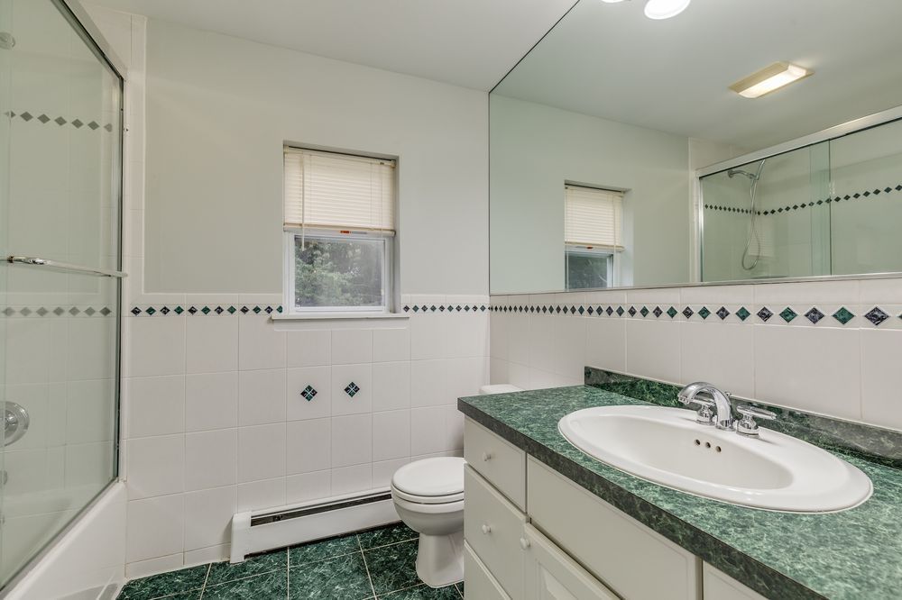 Bathroom with white tile walls, green countertop and floor. Features a white sink, toilet, and shower, with a window and a large mirror.