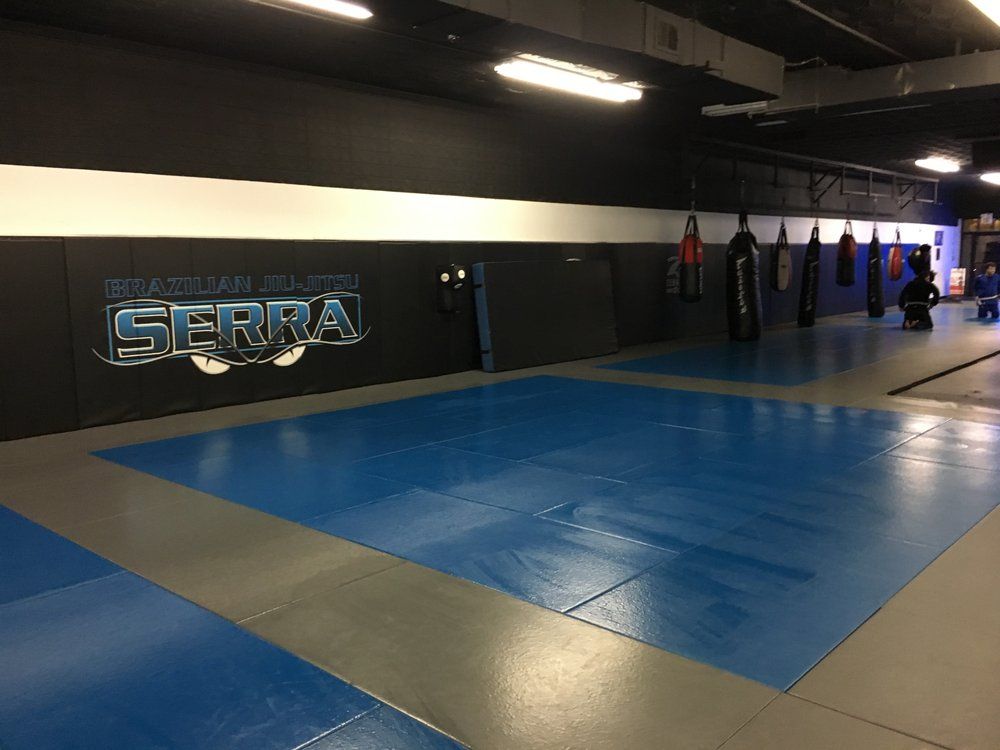Inside a Brazilian Jiu-Jitsu gym, blue and gray mats cover the floor. Black padded walls with a logo line the room, which has hanging heavy bags.