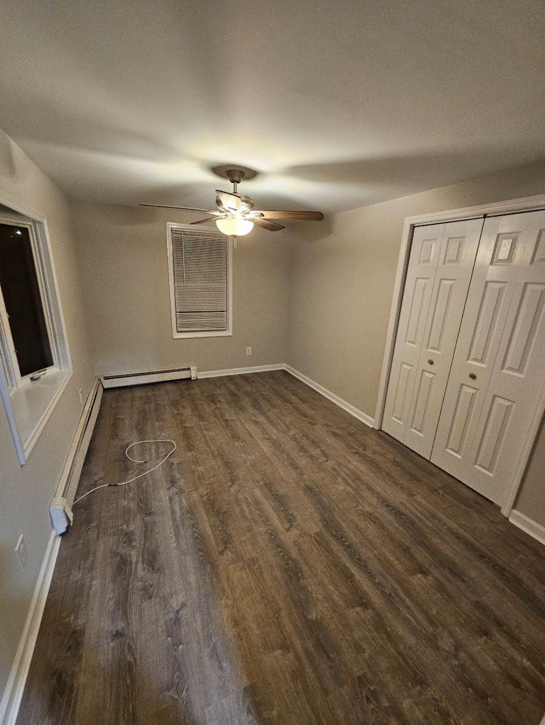 An empty bedroom with wood-look flooring, a ceiling fan, a window, and a closet with white doors. The walls are painted a light beige.