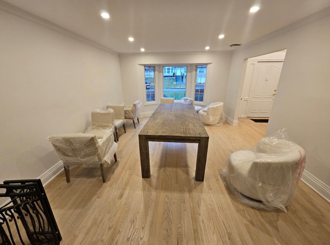 Dining room with long wooden table, six chairs covered in plastic, and a doorway. Neutral walls and light wood floors.
