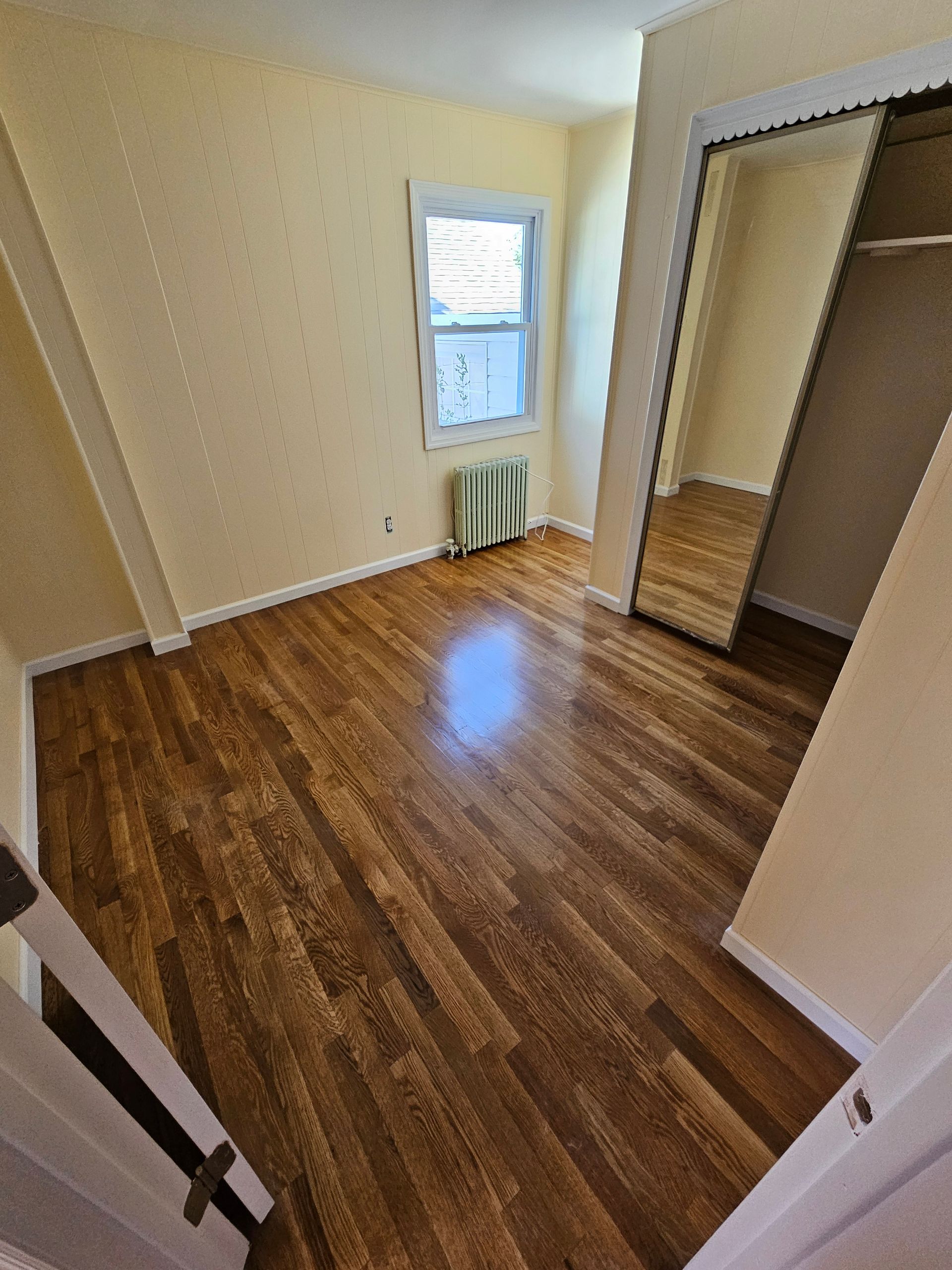 An empty bedroom with hardwood floors, a window with a radiator, and a mirrored closet. Walls are beige.