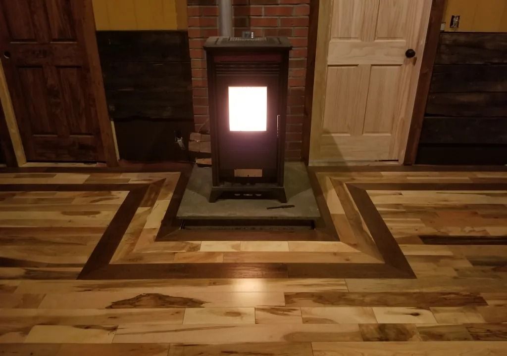 A wood-burning stove with a lit window sits on a dark stone base, centered on a wood floor with a darker border. Doors are visible on each side.