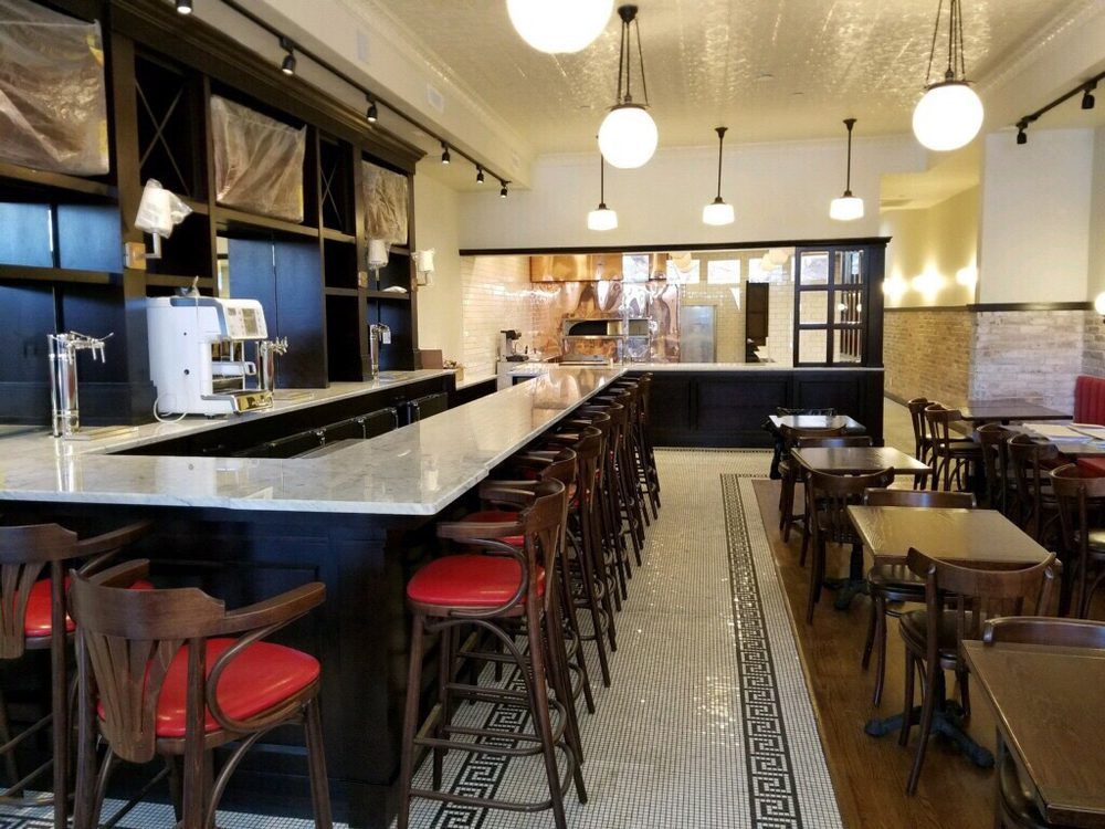 Interior of a restaurant with a long bar, red bar stools, and tables. Tile flooring and hanging globe lights.