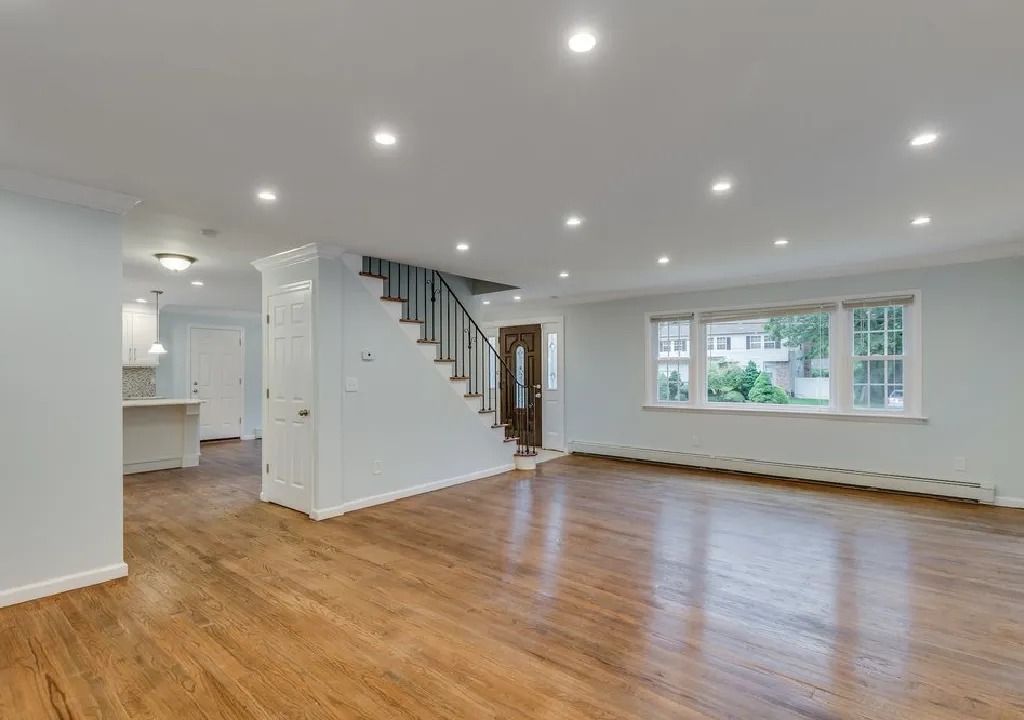 Empty living room with hardwood floors, a staircase, and a window overlooking a green space. The walls are light blue.